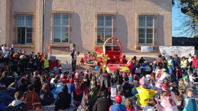 Un gruppo di bambini vestiti in costume da carnevale è seduto a terra a guardare un artista su un palco davanti a Palazzo Doria a Loano. È presente anche un carro allegorico colorato e un uomo in costume.