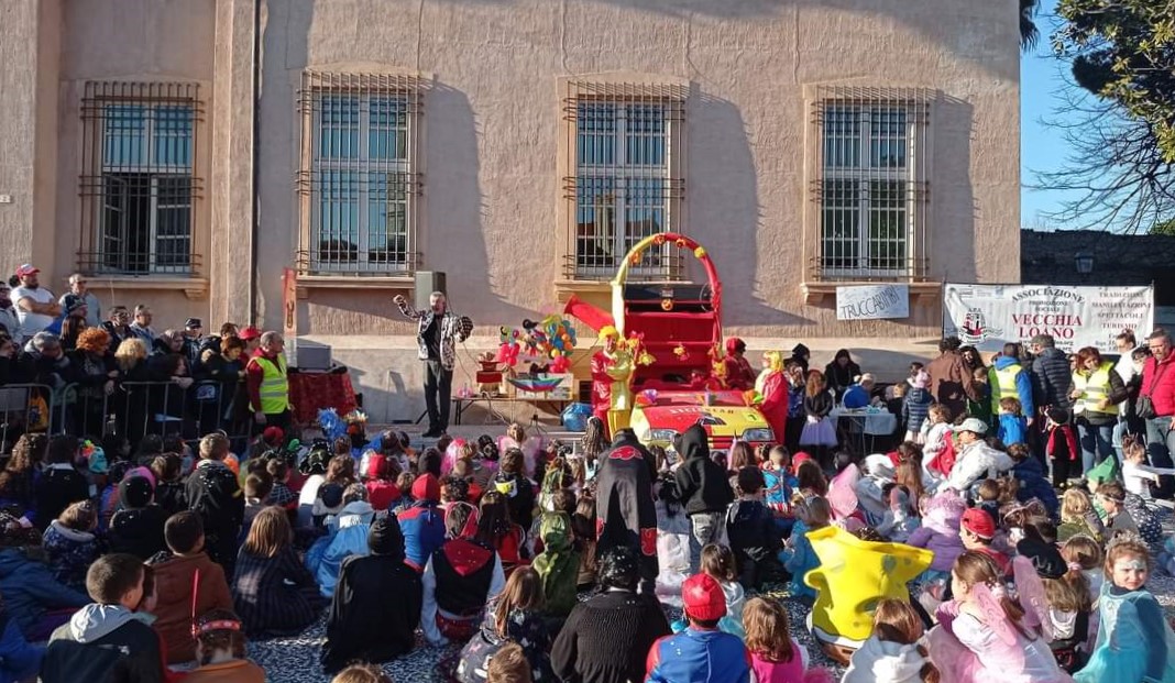 Un gruppo di bambini vestiti in costume da carnevale è seduto a terra a guardare un artista su un palco davanti a Palazzo Doria a Loano. È presente anche un carro allegorico colorato e un uomo in costume.