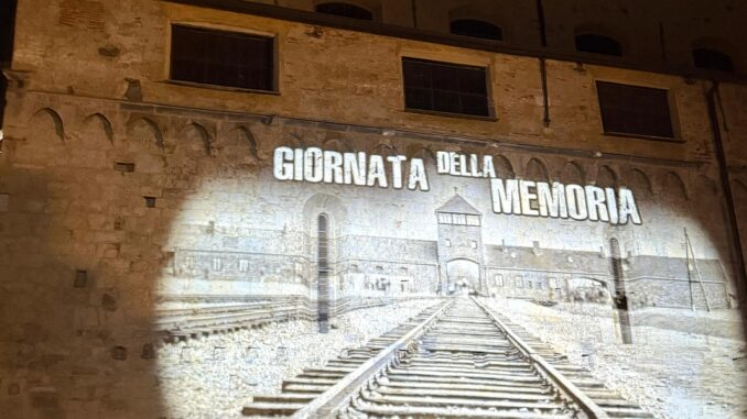 L'immagine mostra una proiezione del campo di concentramento di Auschwitz su un edificio del centro storico di Albenga. Sopra l'immagine è proiettata la frase "Giornata della Memoria".