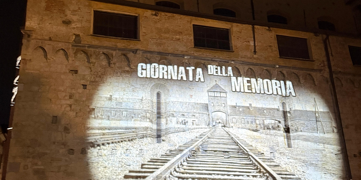 L'immagine mostra una proiezione del campo di concentramento di Auschwitz su un edificio del centro storico di Albenga. Sopra l'immagine è proiettata la frase "Giornata della Memoria".
