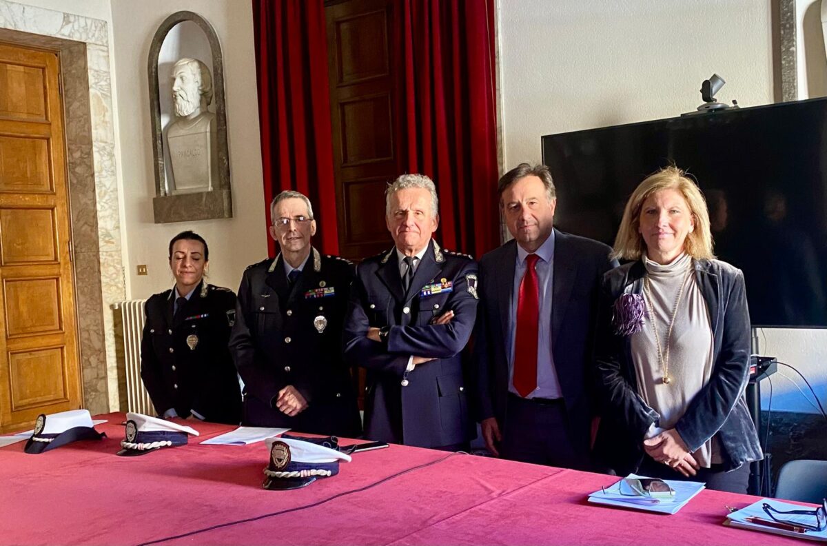 Nella sala del comune un gruppo di cinque persone, tre uomini e due donne, è in piedi dietro a un tavolo coperto da un panno rosso. Diverse cappelli della polizia sono posizionati sul tavolo. Tra loro ci sono il sindaco di Savona Marco Russo, l'assessore alla Polizia Urbana Barbara Pasquali, il comandante della Polizia Locale Igor Aloi e la vicecomandante Francesca Ferba