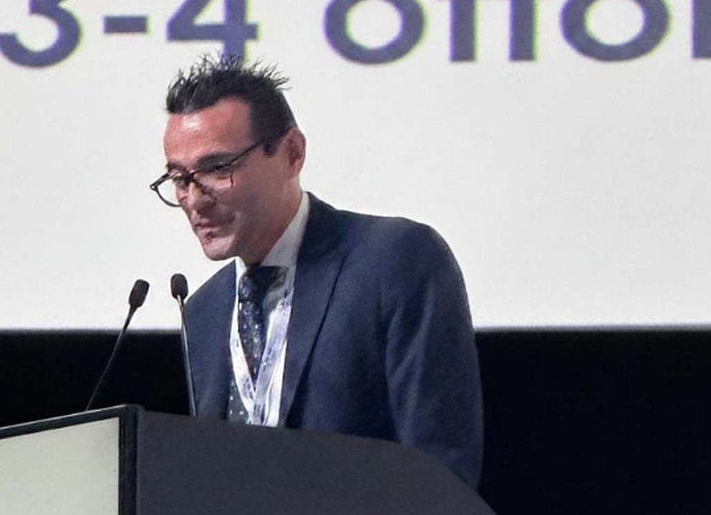 Alessio Bellinghieri della Fiaip in abito e cravatta con gli occhiali è in piedi a un podio con microfoni durante un convegno. Guarda verso il basso e di lato. Sullo sfondo, un grande schermo.