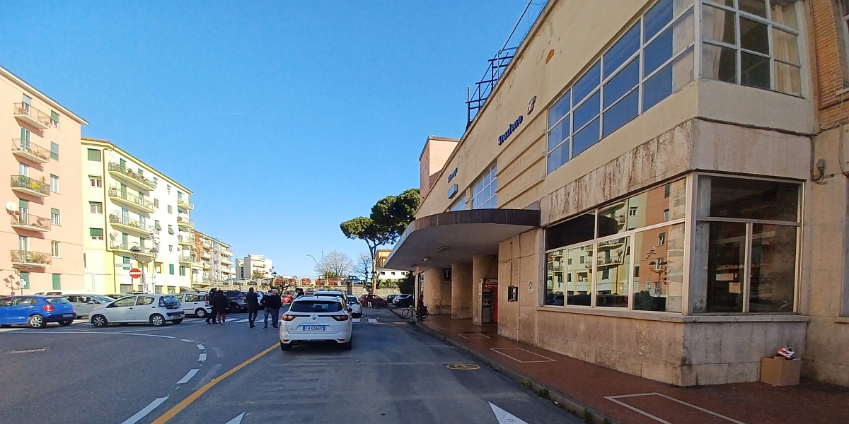 Una scena di strada davanti a un edificio con grandi finestre, la stazione ferroviaria di Albenga, con automobili e palazzi residenziali sullo sfondo. Il cielo è di un azzurro limpido.