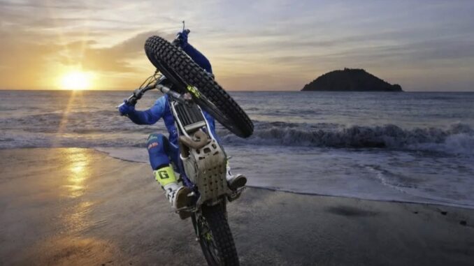 La suggestiva immagine di una moto da trial che si impenna in riva al mare sulla spiaggia di Albenga con sullo sfondo l'Isola Gallinara e il sole che sorge