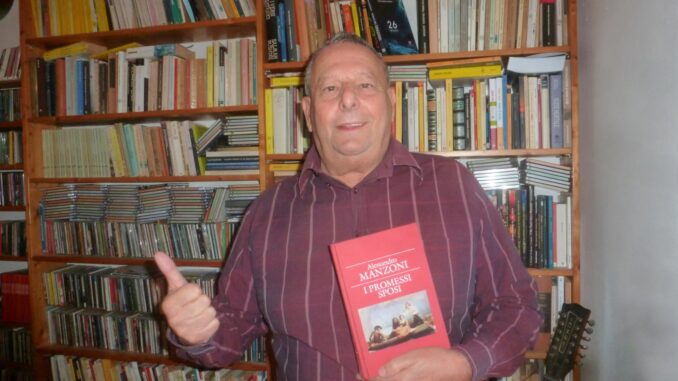 Lo scrittore Maurizio Pupi Bracali davanti agli scaffali di una biblioteca con in mano una copia dei Promessi sposi